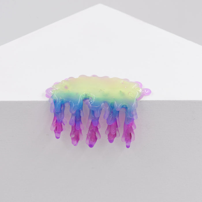 The Mini #7.17 by Dan Lam is a small, abstract resin sculpture with yellow to blue and purple gradients and drip-like extensions, placed on the edge of a white geometric surface against a plain background.