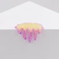 Dan Lams Mini #7.20, a glossy resin sculpture in abstract yellow, pink, and purple hues, resembles melted wax as it drips over the edge of a white triangular surface against a light gray background.