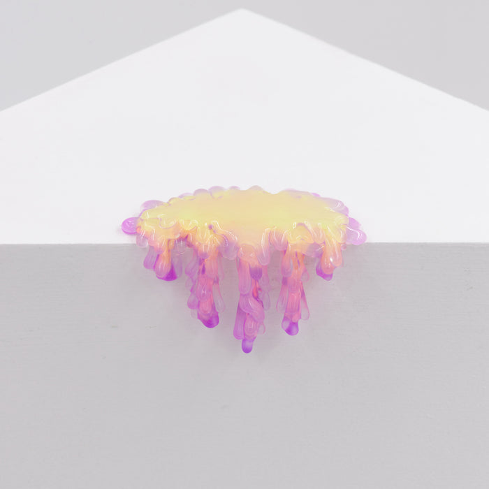 Dan Lams Mini #7.20, a glossy resin sculpture in abstract yellow, pink, and purple hues, resembles melted wax as it drips over the edge of a white triangular surface against a light gray background.