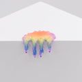 Dan Lam's 'Mini #7.21,' a glossy, rainbow-colored blob resembling poured resin with drips, hangs over the edge of a white geometric surface against a plain light gray background.