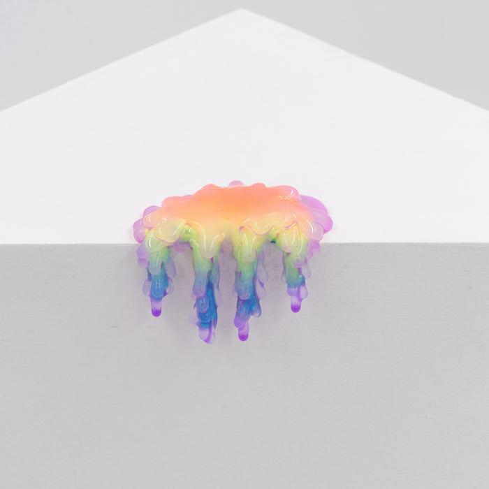 Dan Lam's 'Mini #7.21,' a glossy, rainbow-colored blob resembling poured resin with drips, hangs over the edge of a white geometric surface against a plain light gray background.