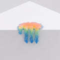 Dan Lams Mini #7.35 is a small, glossy piece resembling melting resin or gel with a blend of orange, yellow, green, and blue colors. It hangs over a white triangular surface set against a plain light gray background.