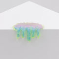 A small, rounded pastel-colored sculpture titled "Mini #7.37" by Dan Lam, featuring glossy pink, blue, and green resin drips, hangs over the edge of a white geometric surface against a light gray background.