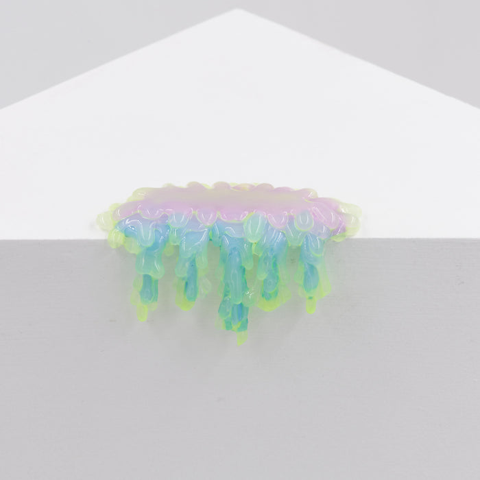 A small, rounded pastel-colored sculpture titled "Mini #7.37" by Dan Lam, featuring glossy pink, blue, and green resin drips, hangs over the edge of a white geometric surface against a light gray background.