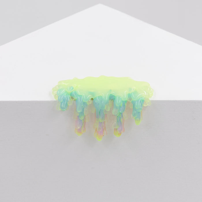 The Dan Lam Mini #7.44 sculpture, an iridescent, drippy resin piece with green, blue, and pink hues, rests elegantly on a white surface like a melting puddle formed by delicate acrylic layers.