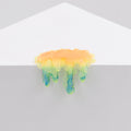 Dan Lam's "Mini #7.45," a glossy, drip-shaped resin object in yellow, green, and blue hues, hangs over the edge of a white geometric surface against a plain light gray background.