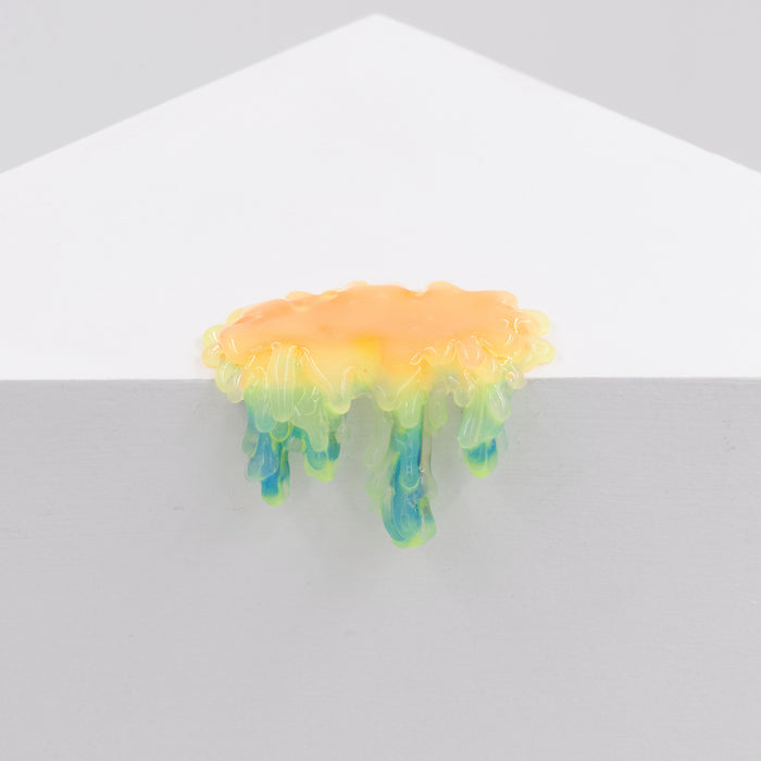 Dan Lam's "Mini #7.45," a glossy, drip-shaped resin object in yellow, green, and blue hues, hangs over the edge of a white geometric surface against a plain light gray background.