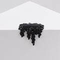 The black enamel pin from Dan Lam, called Mini #7.63, shaped like dripping liquid, elegantly rests on the edge of a white geometric surface, its design mimicking spilled ink or resin and creating a stunning contrast against the minimalistic background.