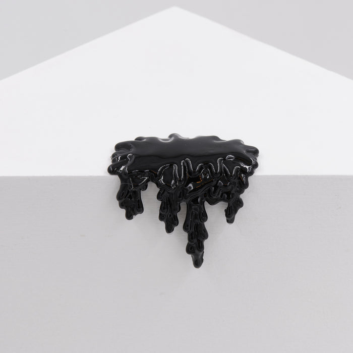 The black enamel pin from Dan Lam, called Mini #7.63, shaped like dripping liquid, elegantly rests on the edge of a white geometric surface, its design mimicking spilled ink or resin and creating a stunning contrast against the minimalistic background.