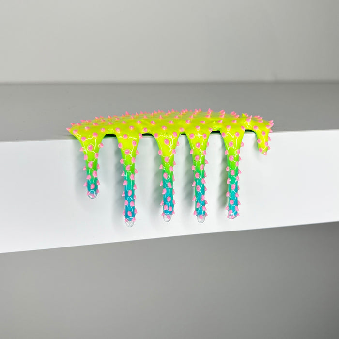 A neon green and blue spiked, goo-like object drips over the edge of a white surface, its form covered in pink-tipped spikes and set against a plain gray background. "Mini #855" by Dan Lam (2023) is a striking mixed media creation that captures otherworldly texture and vibrant energy.