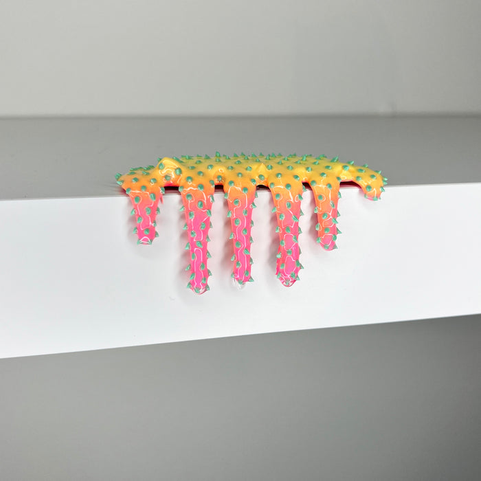 Dan Lam's 'Mini #906'—a yellow and pink spiky sculpture—drips playfully over the edge of a white shelf against a plain gray background, exemplifying the artist's distinctive mixed media style in this 2023 piece.