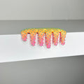 Dan Lam's "Mini #909" hair claw clip—a striking accessory featuring a vibrant gradient of yellow, orange, and pink adorned with small green spikes—rests on the edge of a white shelf against a gray background, perfectly capturing the playful mixed media artistry that defined 2023.