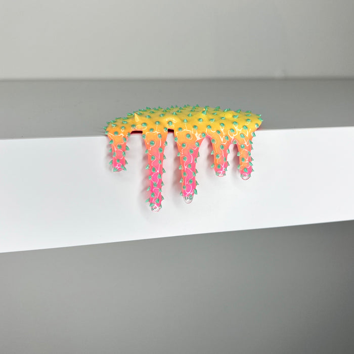 Dan Lam’s "Mini #917" is a yellow and pink spiked mixed media sculpture from 2023, shaped like melting slime as it drips over the edge of a white shelf against a plain gray background.