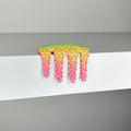Dan Lams Mini #926, a colorful, drippy sculpture from 2023, features spikes and a shiny textured gradient from orange to pink. Made with mixed media, it sits on the edge of a white shelf and measures 3/4 x 2 1/2 x 1 3/4 inches.