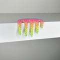 Dan Lam - "Mini #967" by Dan Lam is a colorful, spiky, drippy mixed media sculpture featuring pink, orange, yellow, blue, and green hues. The piece sits on the edge of a white shelf against a plain gray background and was created in 2023.