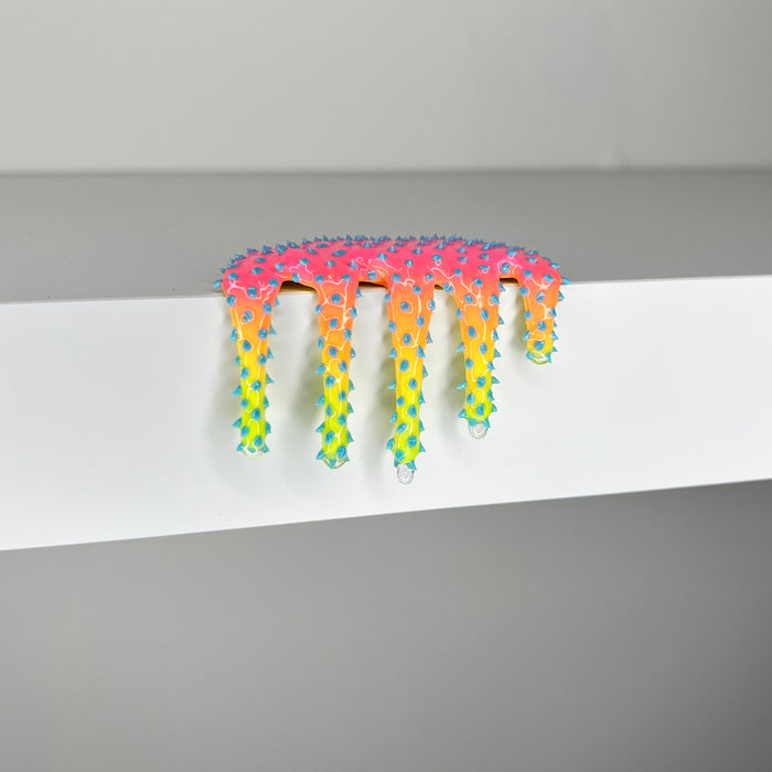 Dan Lam - "Mini #967" by Dan Lam is a colorful, spiky, drippy mixed media sculpture featuring pink, orange, yellow, blue, and green hues. The piece sits on the edge of a white shelf against a plain gray background and was created in 2023.