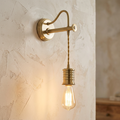 Aged brass industrial wall light with a curved arm and hanging Edison bulb against a textured plaster wall.