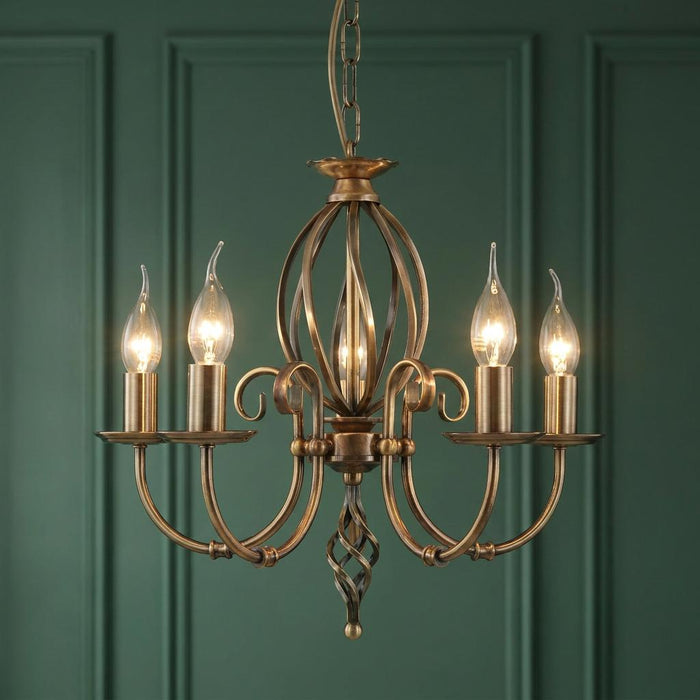 Aged brass 5-light chandelier with flame-shaped bulbs against a dark green paneled wall.