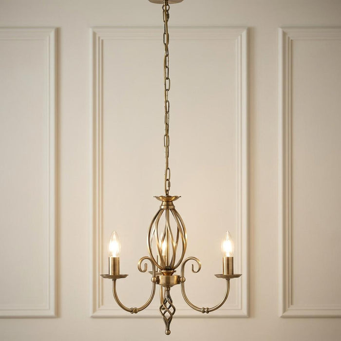 Elstead Artisan 3-light aged brass chandelier with scroll arms and birdcage detail against a paneled wall.