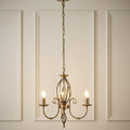 Elstead Artisan 3-light aged brass chandelier with scroll arms and birdcage detail against a paneled wall.