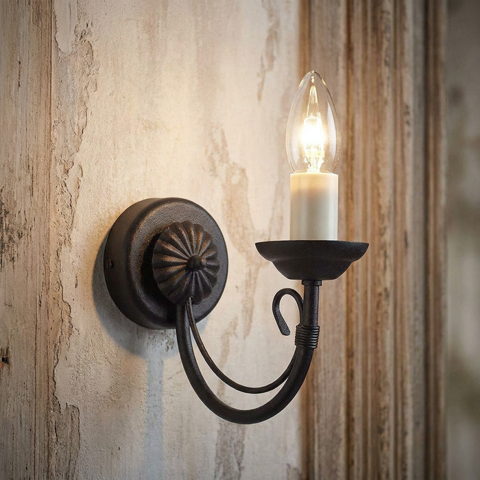 Black wrought iron wall light with a glowing candle-style bulb mounted on a rustic textured wall.