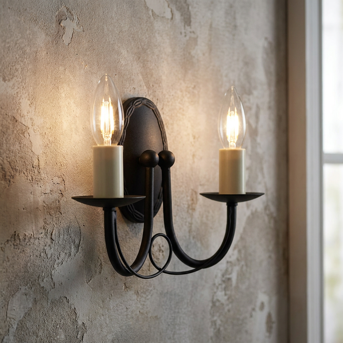 Elstead Minster 2-light black wall sconce with glowing candle-style bulbs on a rustic, textured wall.