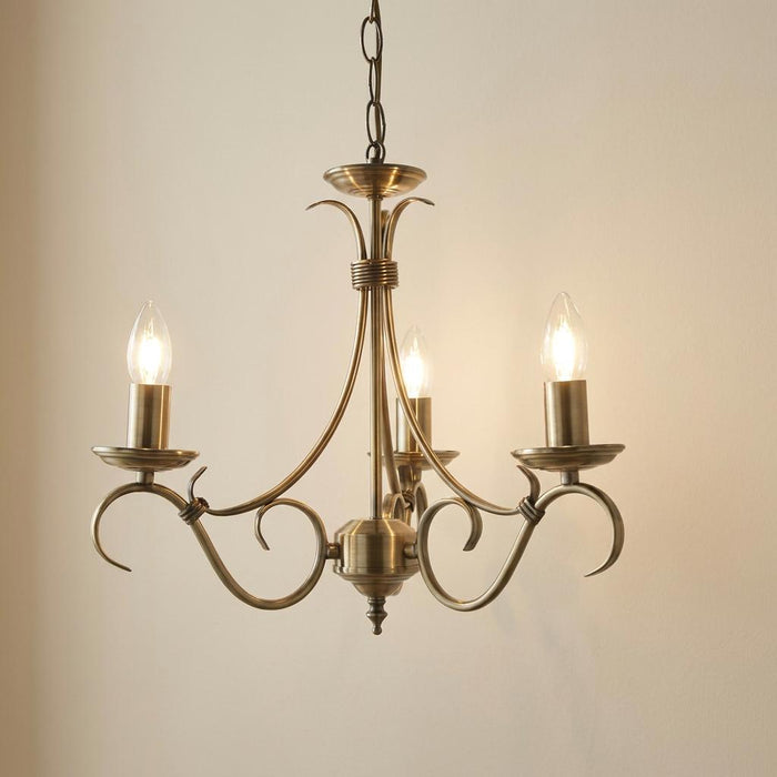 Endon Bernice antique brass 3-light chandelier with decorative curved arms and glowing candle bulbs against a plain wall.