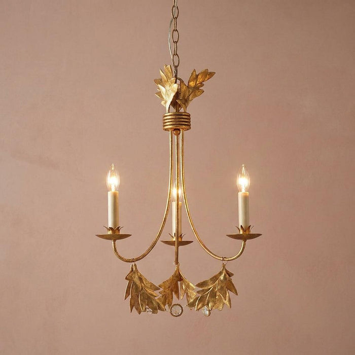 Three-light gilded gold chandelier with olive leaf motifs and lit candle bulbs against a plain pink wall.
