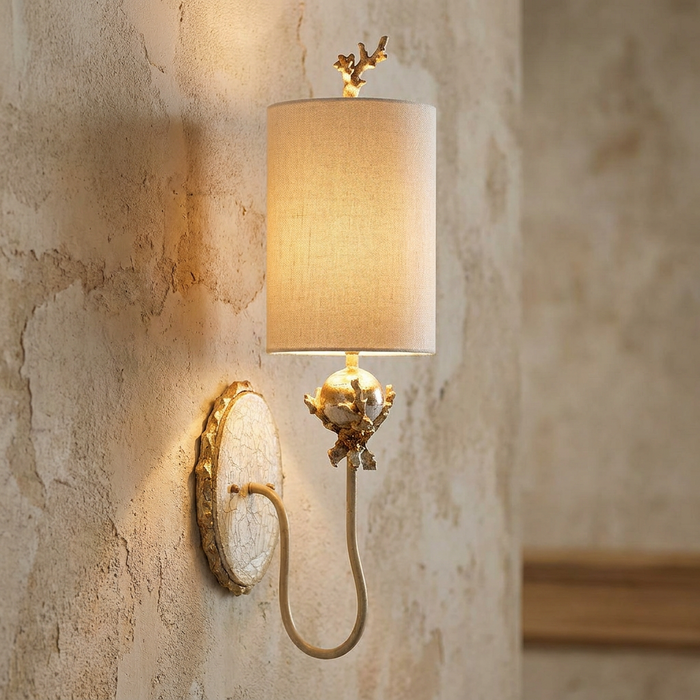 Putty and silver wall light with coral-like details and a fabric shade, glowing against a textured wall.