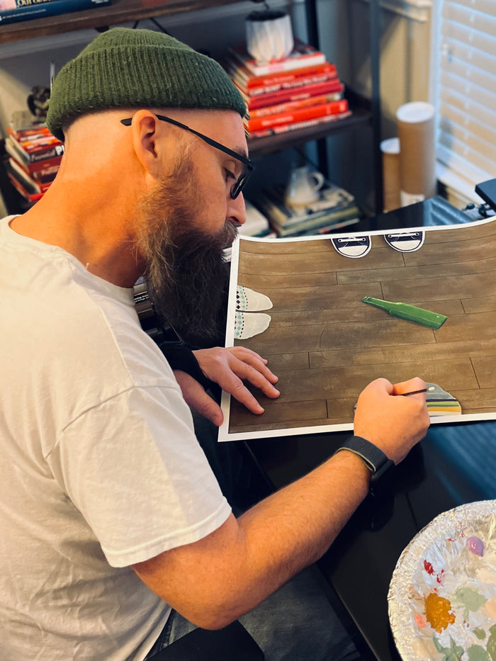 A man wearing a green beanie and glasses sketches at a desk surrounded by art supplies, books, and a palette. He is creating "Spin," a limited edition archival pigment print by Gabe Langholtz, which features details such as a wooden floor, shoes, and a green bottle.