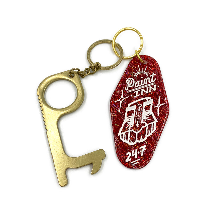 The GATS - "Paint INN 24.7" Keychain by GATS features a brass multi-purpose tool attached to an acrylic keychain with a red, vintage motel-style tag reading "GATS Paint INN," adorned with an illustration of two hands and the numbers "24-7" below.