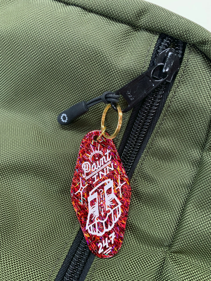 A close-up of a green bag with a black zipper, featuring the GATS - "Paint INN 24.7" Keychain by GATS—a red acrylic accessory styled like a vintage motel keychain, adorned with “Paint Inn 24/7” text and an illustration of two hands with white branches in the background, paying tribute to the iconic GATS Paint INN collection.
