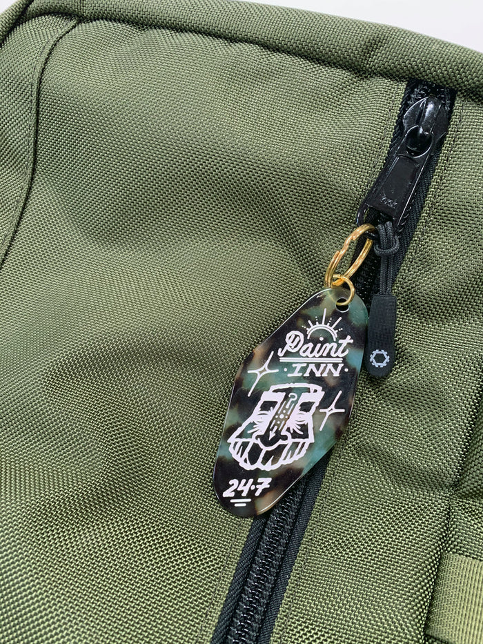 A green textured bag with a black zipper, featuring a GATS "Paint INN 24.7" keychain attached—a vintage motel-inspired acrylic accessory from GATS, showcasing a white skull and crossbones on a black and green background that channels the brand's edgy Paint INN style.