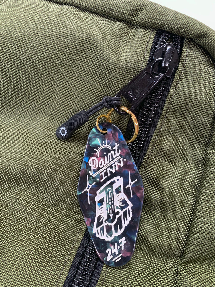 A green fabric bag with a zippered pocket is adorned with a GATS - "Paint INN 24.7" Keychain, which features the brand's stylized face drawing and text "Paint INN 24.7" in white on a marbled, dark acrylic background, reminiscent of vintage motel keychains.