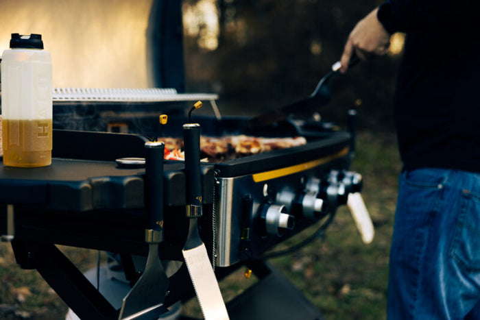 Black gas grill with yellow accents, multiple burners, cooking food on grates with person grilling