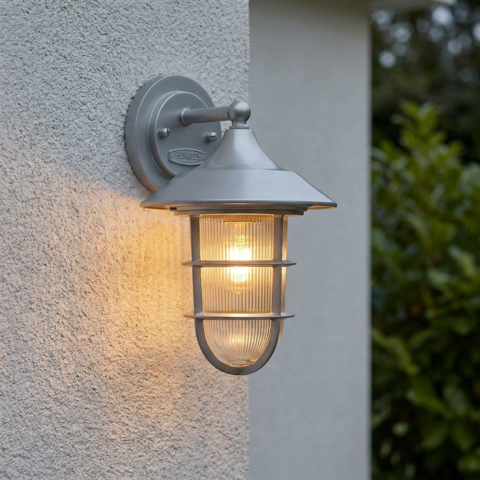 Silver nautical-style wall lantern with ribbed glass and a warm glow mounted on a textured white exterior wall.