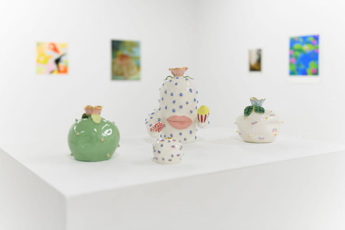 Four small whimsical ceramic sculptures by Jen Dwyer titled Harriet, featuring abstract designs with pastel colors and gold luster, are displayed on a white pedestal in a gallery. Blurred colorful paintings adorn the walls in the background.