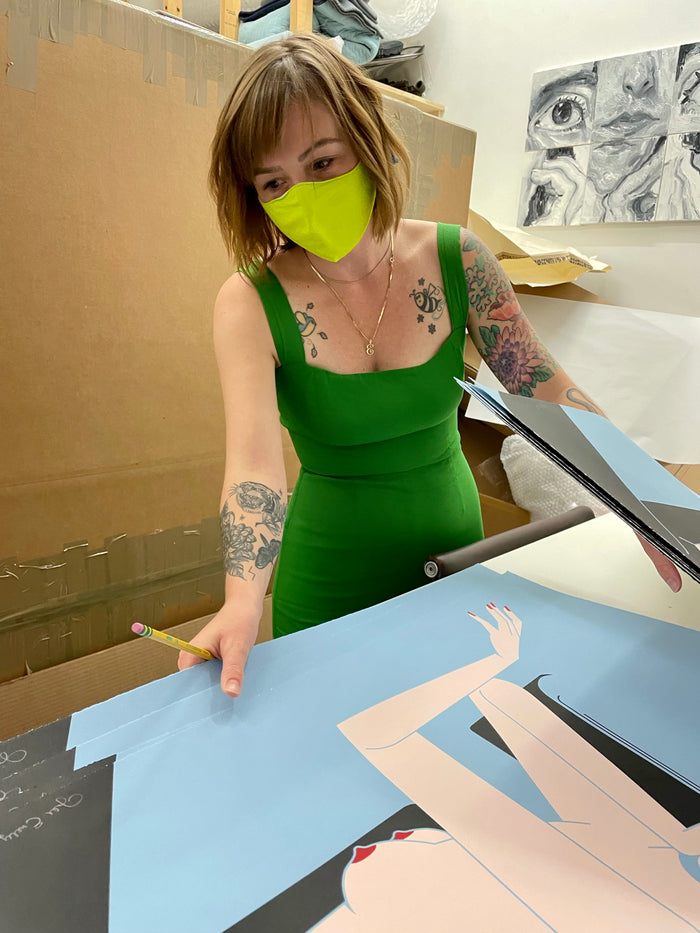 A woman in a green dress and yellow mask, with tattoos on her arms, points at the Jillian Evelyn - "Attempt" limited edition screenprint displayed on Coventry Rag, which is laid out on a table in a studio filled with cardboard boxes and art supplies in the background.