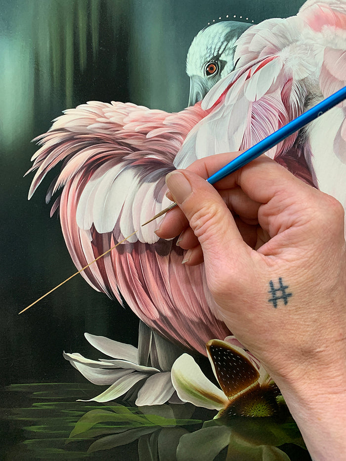 A hand holding a thin paintbrush carefully paints intricate details on a pink bird with soft feathers, while a hashtag tattoo is visible on the artist's hand. "Watershed" by Josie Morway is offered as a signed and numbered archival pigment print in a limited edition.