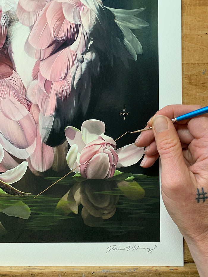 A tattooed hand holds a small paintbrush, carefully adding details to Josie Morway’s "Watershed" print—a realistic portrayal of pink feathers and white flowers reflected on water; this limited edition artwork from Josie Morway is signed and numbered at the bottom.