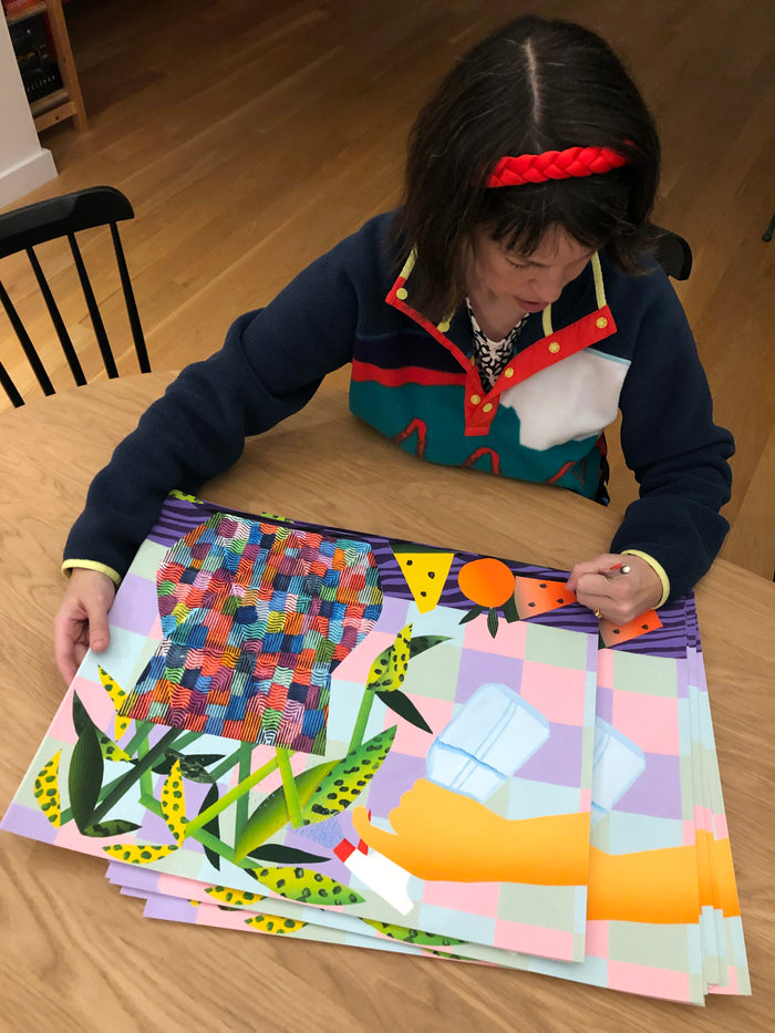 A person with a red headband sits at a wooden table, admiring Karen Lederer's "Spray" print—a limited edition archival pigment print featuring colorful geometric designs of abstract shapes, plants, and fruit set against a checkered background. The artwork’s hand deckled edges and exceptional quality showcase both its artistry and exclusivity.