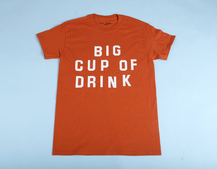 A Katie Kimmel "Big Cup of Drink" short-sleeve cotton t-shirt in orange, featuring large white lettering on the front, is laid flat on a light blue background.