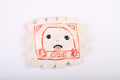 The Katie Kimmel Stamp (red) is a whimsical ceramic piece in a scalloped square shape, featuring a cartoon face with big eyes and a cheeky tongue. The minimalistic design is framed by the word ONE and the number 1, making it charming and unique.