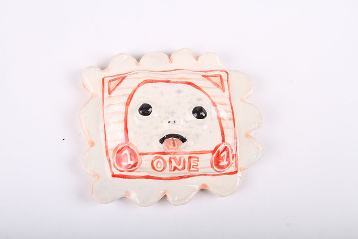The Katie Kimmel Stamp (red) is a whimsical ceramic piece in a scalloped square shape, featuring a cartoon face with big eyes and a cheeky tongue. The minimalistic design is framed by the word ONE and the number 1, making it charming and unique.