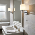 Chrome bathroom wall light with a glowing opal glass shade mounted on a tiled wall beside a large mirror.