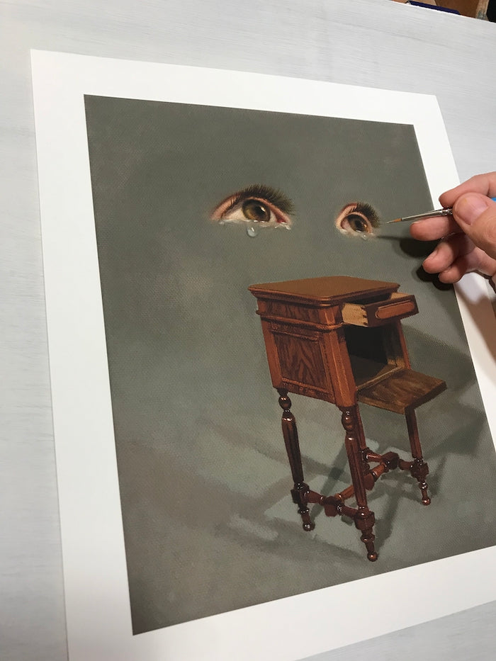 A hand with a paintbrush adds details to Lola Gils Open Conversation print, where a small wooden table with an open drawer stands beneath two realistic eyes with teardrops, crafting a dreamlike scene. This surreal art piece is part of Lola Gils signed and numbered edition collection.