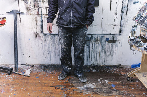 In an art studio, a person in paint-splattered clothes stands among vibrant drips and splatters. Near the easel holding Shaun Roberts custom-framed Psyche with Brett Amory, Oakland, CA, scattered tools lie around. The artists face blends into the creative chaos.