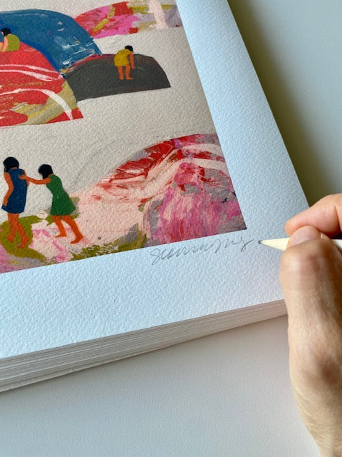 Hand holding pencil signing "Seonna Hong" on print of people standing and climbing onto rainbow colored rocks over a beige background