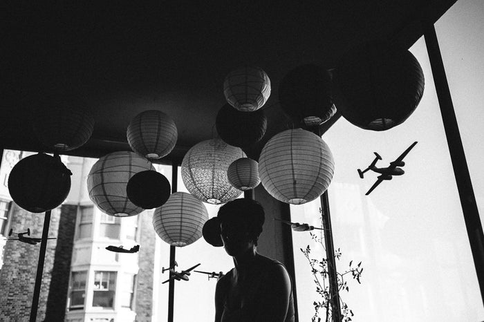 A silhouette stands by large windows amidst hanging paper lanterns and model airplanes. Buildings loom beyond in this black-and-white scene, captured in a signed and numbered archival pigment print titled “Untitled” (with Jessica Hess, San Francisco, CA), by Shaun Roberts.